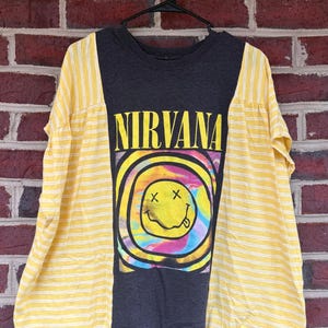 May include: A dark gray t-shirt with the word "NIRVANA" in yellow block letters. The shirt features a colorful smiley face design. The sleeves are yellow with white vertical stripes. The shirt is hanging on a black hanger.