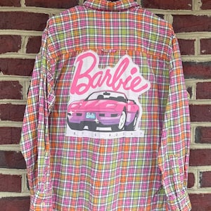 May include: A button-down shirt with a colorful plaid pattern in pink, orange, green, and yellow. The back features a large graphic of the Barbie logo and a pink convertible car. The text "LET'S ROLL" is below the car.