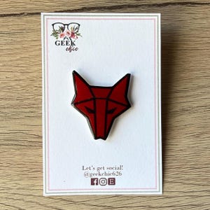 May include: A red and black geometric fox head enamel pin attached to a white display card. The card features the text GEEK chic, Let's get social! @geekchic626, and icons for Facebook, Instagram, and Etsy. The fox design has sharp, angular lines.