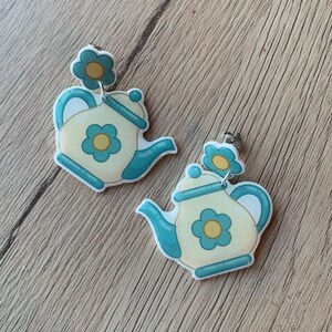 May include: A pair of blue and white teapot earrings with yellow flower accents. The earrings are made of polymer clay and have silver hooks.