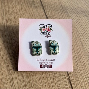 May include: A pair of white and blue Star Wars Clone Trooper helmet earrings. The earrings are on a pink card with the text "Geek chic" and social media icons.