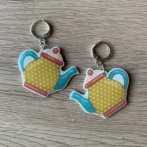 May include: A pair of earrings featuring a yellow teapot with blue and white accents. The teapot has a polka dot pattern and is attached to a silver hoop.