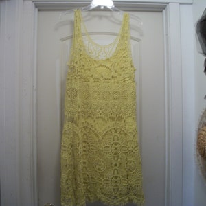 Puede incluir: Vestido de crochet amarillo p&aacute;lido con dise&ntilde;o sin mangas, colgado de una percha de pl&aacute;stico transparente. El vestido presenta intrincados patrones calados y un dobladillo con flecos, lo que sugiere un estilo bohemio o playero.