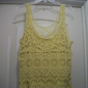Puede incluir: Camiseta sin mangas de crochet amarillo p&aacute;lido con un delicado estampado floral y detalles de flecos. La parte superior est&aacute; colgada de una percha de pl&aacute;stico transparente contra una puerta blanca. La camiseta sin mangas est&aacute; hecha de un material suave y ligero.