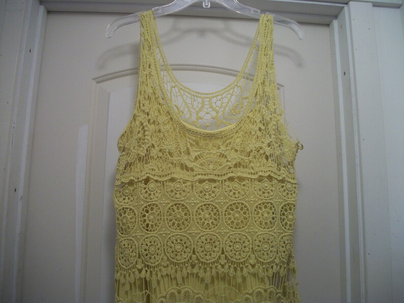 Puede incluir: Camiseta sin mangas de crochet amarillo p&aacute;lido con un delicado estampado floral y detalles de flecos. La parte superior est&aacute; colgada de una percha de pl&aacute;stico transparente contra una puerta blanca. La camiseta sin mangas est&aacute; hecha de un material suave y ligero.