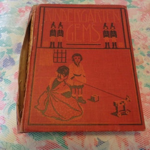 May include: A vintage book titled "Kindergarten Gems" with a red cover and illustrations. The cover features children and the title in a decorative font. The book's spine shows wear, indicating its age.