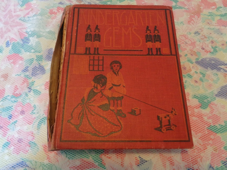 May include: A vintage book titled "Kindergarten Gems" with a red cover and illustrations. The cover features children and the title in a decorative font. The book's spine shows wear, indicating its age.