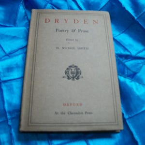 May include: A vintage book titled "Dryden Poetry & Prose" with a beige cover and red lettering. The book is edited by D. Nichol Smith and published by Oxford at the Clarendon Press. The book is on a blue fabric surface.