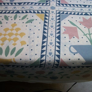 May include: White fabric with a repeating pattern of floral and geometric designs. The pattern includes stylized flowers, leaves, and geometric shapes in yellow, pink, blue, and green. The border features blue triangles and hearts.
