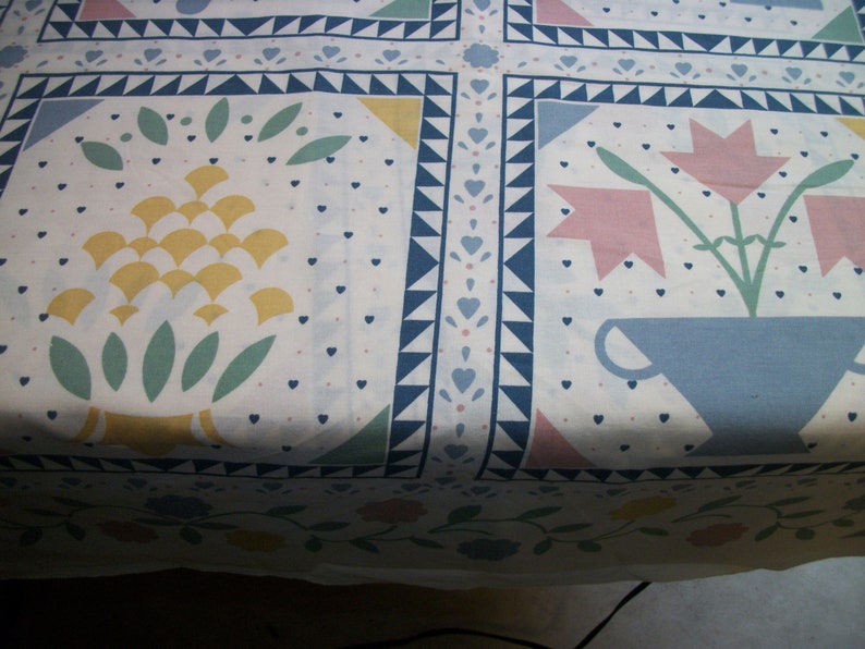 May include: White fabric with a repeating pattern of floral and geometric designs. The pattern includes stylized flowers, leaves, and geometric shapes in yellow, pink, blue, and green. The border features blue triangles and hearts.