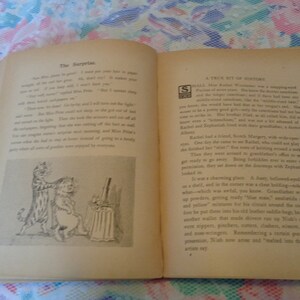 May include: An open vintage book with red binding, featuring printed text and a black and white illustration of two cats. The pages are aged with a warm, yellowed tone, suggesting the book's age. The text includes the title "The Surprise."