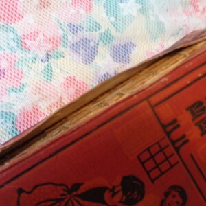 May include: Close-up of a vintage book with a red cover and illustrations. The book's spine is damaged, revealing the aged pages. The background features a floral patterned fabric in pink, blue, and white.