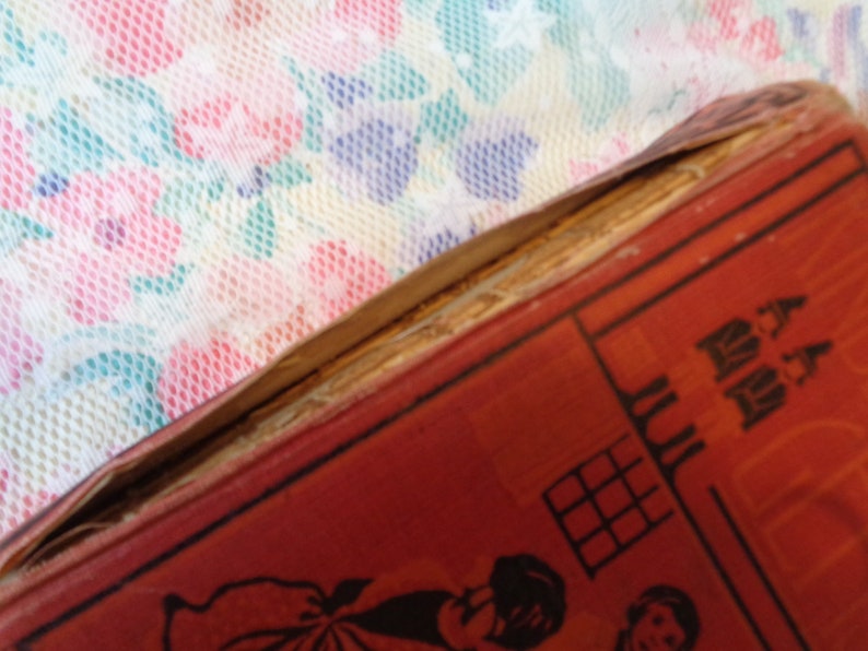 May include: Close-up of a vintage book with a red cover and illustrations. The book's spine is damaged, revealing the aged pages. The background features a floral patterned fabric in pink, blue, and white.