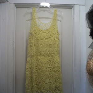 Puede incluir: Un vestido amarillo de ganchillo con cuello redondo y dise&ntilde;o sin mangas cuelga de una percha de pl&aacute;stico transparente. El vestido presenta intrincados patrones florales y geom&eacute;tricos. El vestido est&aacute; hecho de un material suave y ligero.