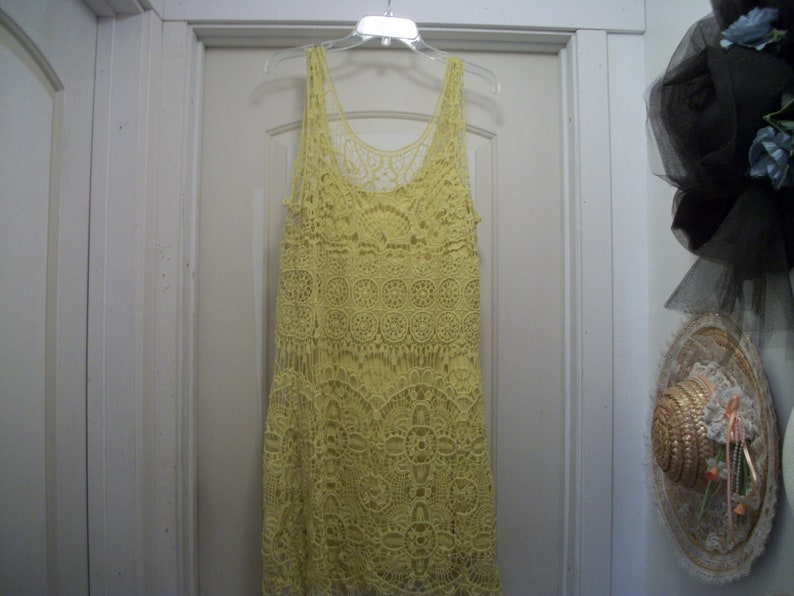 Puede incluir: Un vestido amarillo de ganchillo con cuello redondo y dise&ntilde;o sin mangas cuelga de una percha de pl&aacute;stico transparente. El vestido presenta intrincados patrones florales y geom&eacute;tricos. El vestido est&aacute; hecho de un material suave y ligero.