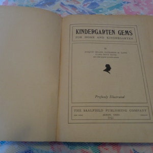 May include: An open book with the title "KINDERGARTEN GEMS" on the right page. The cover is a light tan color with black text and a silhouette. The book is "Profusely Illustrated" and published by The Saalfield Publishing Company.