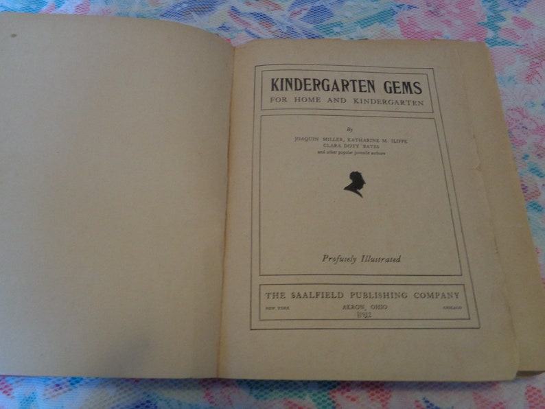 May include: An open book with the title "KINDERGARTEN GEMS" on the right page. The cover is a light tan color with black text and a silhouette. The book is "Profusely Illustrated" and published by The Saalfield Publishing Company.