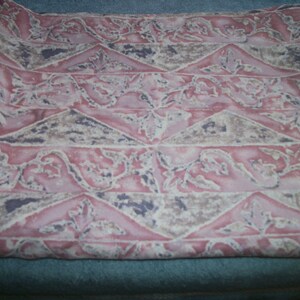 May include: A folded textile featuring a repeating pattern of pink, beige, and purple geometric shapes and floral elements. The fabric has a soft, slightly wrinkled texture. The design is symmetrical and decorative, suitable for various uses.