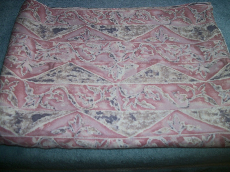 May include: A folded textile featuring a repeating pattern of pink, beige, and purple geometric shapes and floral elements. The fabric has a soft, slightly wrinkled texture. The design is symmetrical and decorative, suitable for various uses.