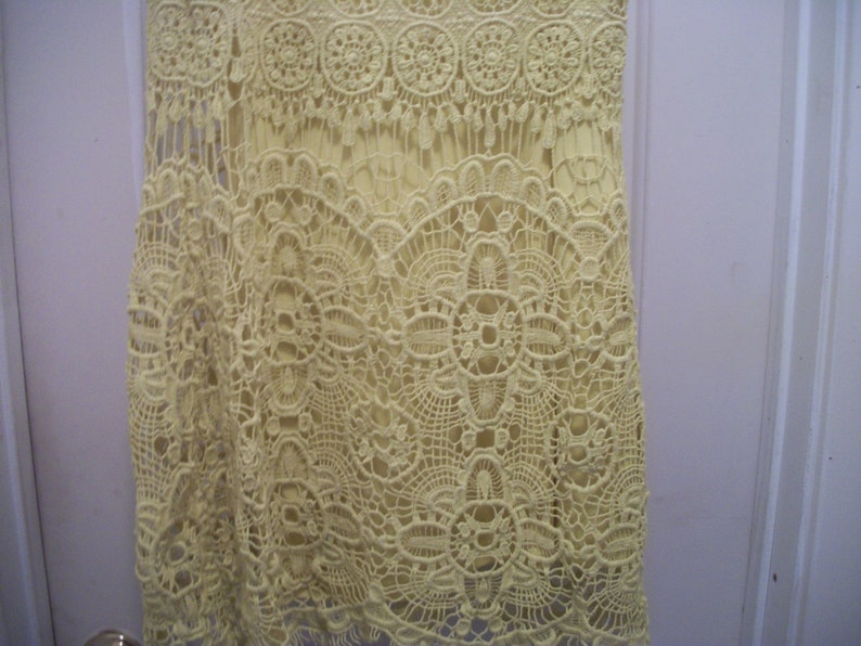 Puede incluir: Primer plano de un vestido amarillo p&aacute;lido con intrincados detalles de encaje de ganchillo. El vestido presenta un dise&ntilde;o en capas con un delicado patr&oacute;n calado. El trabajo de ganchillo crea una apariencia texturizada y aireada, adecuada para el verano.