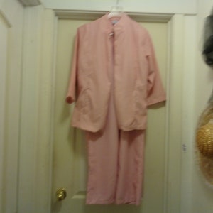 May include: A pale pink two-piece suit, comprising a jacket and trousers, hangs on a hanger. The jacket features three-quarter sleeves and two front pockets. The trousers are straight-legged. The suit is made of a lightweight fabric.