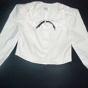 May include: A white cropped jacket with a lace collar and a black satin bow. The jacket has long sleeves and a button closure.