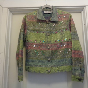 May include: A green, pink and blue striped denim jacket with colourful rhinestones. The jacket has a button closure and two pockets.