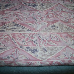 May include: Close-up of a fabric featuring a repeating pattern in pink, beige, and dark grey. The design incorporates triangles and floral motifs, giving a textured appearance. The fabric has a soft, faded aesthetic.