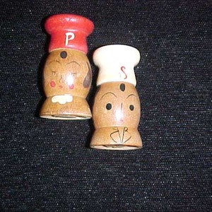 May include: Two wooden salt and pepper shakers with painted faces. The salt shaker has a red and white chef's hat and the pepper shaker has a white chef's hat.