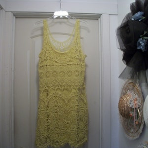 Puede incluir: Un vestido amarillo de ganchillo con dise&ntilde;o sin mangas cuelga de una percha. El vestido presenta un patr&oacute;n calado detallado y un escote redondo. Otros sombreros son visibles en el fondo.