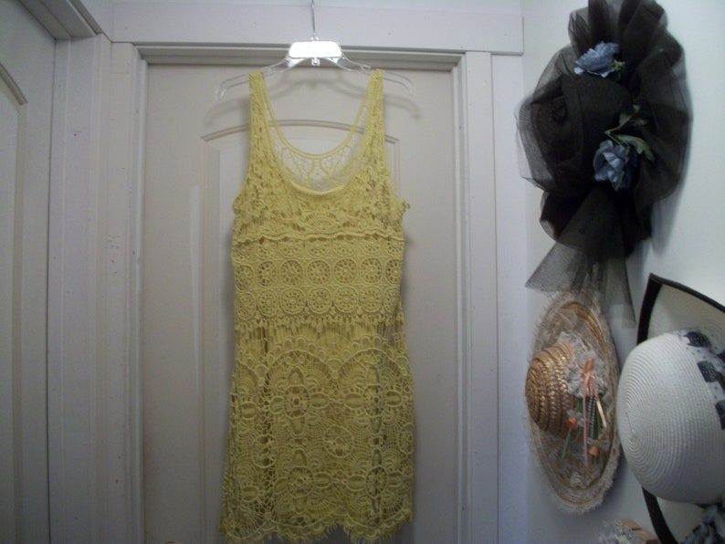 Puede incluir: Un vestido amarillo de ganchillo con dise&ntilde;o sin mangas cuelga de una percha. El vestido presenta un patr&oacute;n calado detallado y un escote redondo. Otros sombreros son visibles en el fondo.