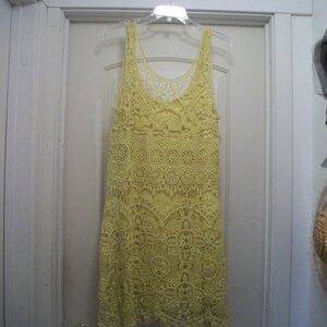 Puede incluir: Un vestido de tirantes de ganchillo amarillo p&aacute;lido cuelga de una percha de pl&aacute;stico transparente. El vestido presenta un cuello redondo y un patr&oacute;n calado detallado. El vestido est&aacute; hecho de un material suave y ligero.