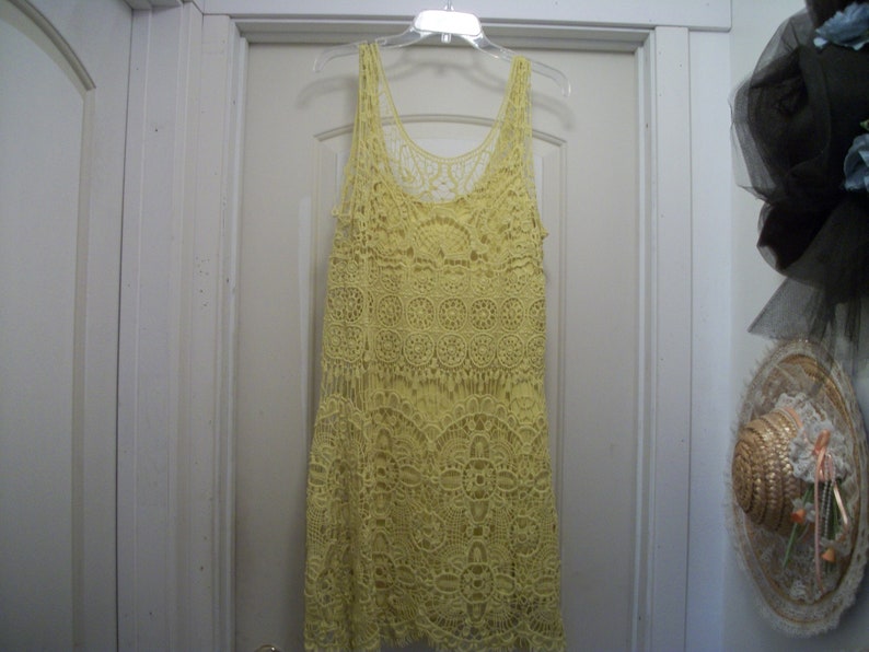 Puede incluir: Un vestido de tirantes de ganchillo amarillo p&aacute;lido cuelga de una percha de pl&aacute;stico transparente. El vestido presenta un cuello redondo y un patr&oacute;n calado detallado. El vestido est&aacute; hecho de un material suave y ligero.