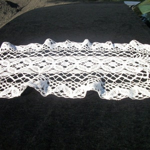 May include: A white, open-weave lace table runner with a scalloped edge. The runner features a repeating floral pattern in the center and is displayed on a dark surface. This decorative piece adds a touch of elegance to any table setting.