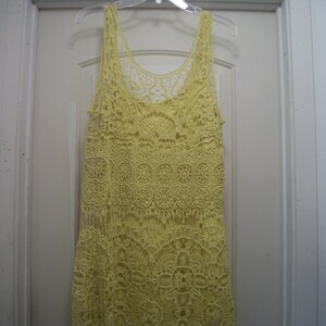 Puede incluir: Un top sin mangas de crochet amarillo con cuello redondo y detalles de flecos. El top est&aacute; hecho de una delicada tela de crochet de tejido abierto. La prenda est&aacute; colgada de una percha de pl&aacute;stico transparente contra una puerta blanca.