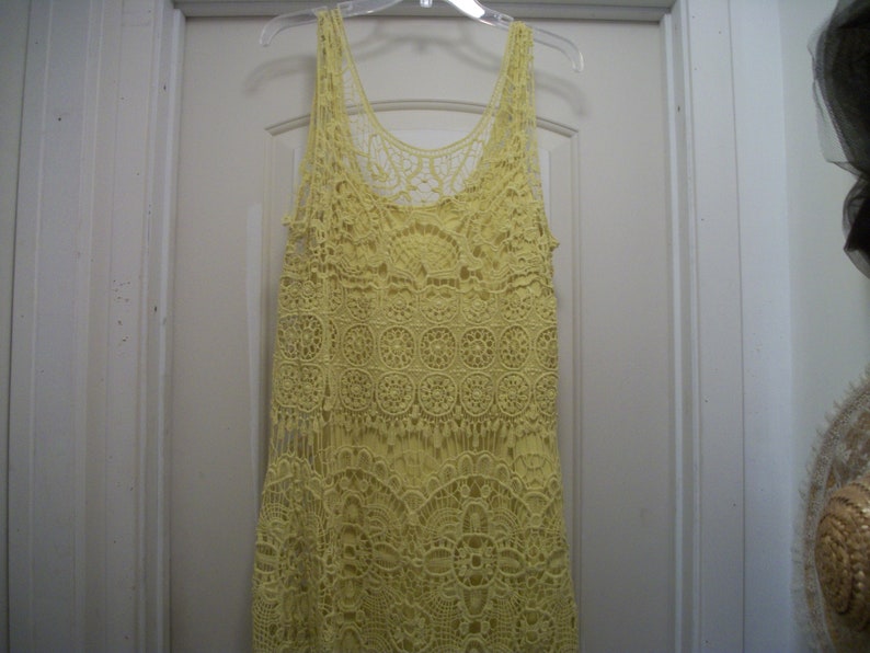 Puede incluir: Un top sin mangas de crochet amarillo con cuello redondo y detalles de flecos. El top est&aacute; hecho de una delicada tela de crochet de tejido abierto. La prenda est&aacute; colgada de una percha de pl&aacute;stico transparente contra una puerta blanca.