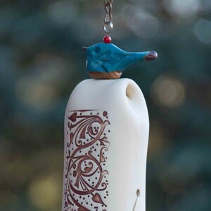 May include: White ceramic wind chime with a blue bird on top and a brown floral design. The chime has a brown bead and a blue bead hanging from the bottom.