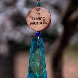 May include: A wooden memorial ornament with the text "In Loving Memory" and a heart symbol. The ornament is attached to a blue and green metal wind chime.