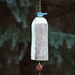 May include: A ceramic wind chime with a white body, adorned with intricate brown patterns and a small blue bird on top. A wooden pendant with a paw print and the inscription "In Memory of Collie" hangs below.