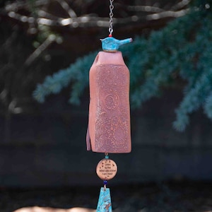 May include: A terracotta wind chime with a blue bird on top and a copper-colored triangle at the bottom. The chime has a wooden tag that reads "Listen to the Wind and Know I am Near".
