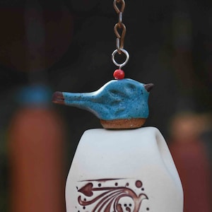 May include: A ceramic wind chime featuring a blue bird-shaped topper with a brown beak, a small red bead, and a metal chain. The white bell body below displays a brown decorative pattern with curved lines and leaf-like shapes on its surface.