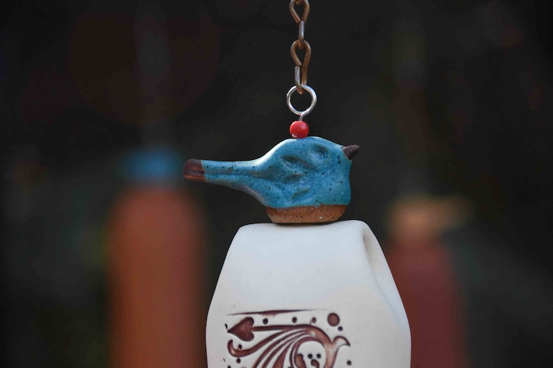 May include: A ceramic wind chime featuring a blue bird-shaped topper with a brown beak, a small red bead, and a metal chain. The white bell body below displays a brown decorative pattern with curved lines and leaf-like shapes on its surface.