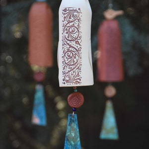 May include: Three hanging ceramic wind chimes featuring a central off-white bell with intricate brown scrollwork patterns. Each chime is topped with a small bird figure and includes a terracotta bead and a dangling, textured blue-green metal pendant.