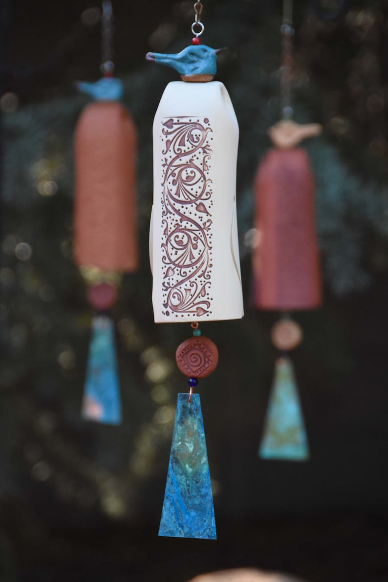 May include: Three hanging ceramic wind chimes featuring a central off-white bell with intricate brown scrollwork patterns. Each chime is topped with a small bird figure and includes a terracotta bead and a dangling, textured blue-green metal pendant.