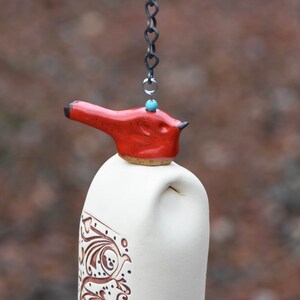 May include: A ceramic wind chime featuring a cream-colored cylindrical body with brown etched scrollwork patterns. A bright red bird-shaped topper sits atop the cylinder, accented by a small turquoise bead and suspended by a dark metal chain.