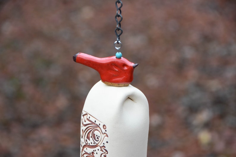 May include: A ceramic wind chime featuring a cream-colored cylindrical body with brown etched scrollwork patterns. A bright red bird-shaped topper sits atop the cylinder, accented by a small turquoise bead and suspended by a dark metal chain.