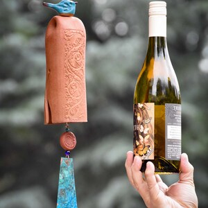 May include: A terracotta wind chime featuring a blue ceramic bird topper, a cylindrical body with embossed floral patterns, a small round terracotta bead, and a flat, triangular copper-toned metal pendant with a blue and green patina finish.