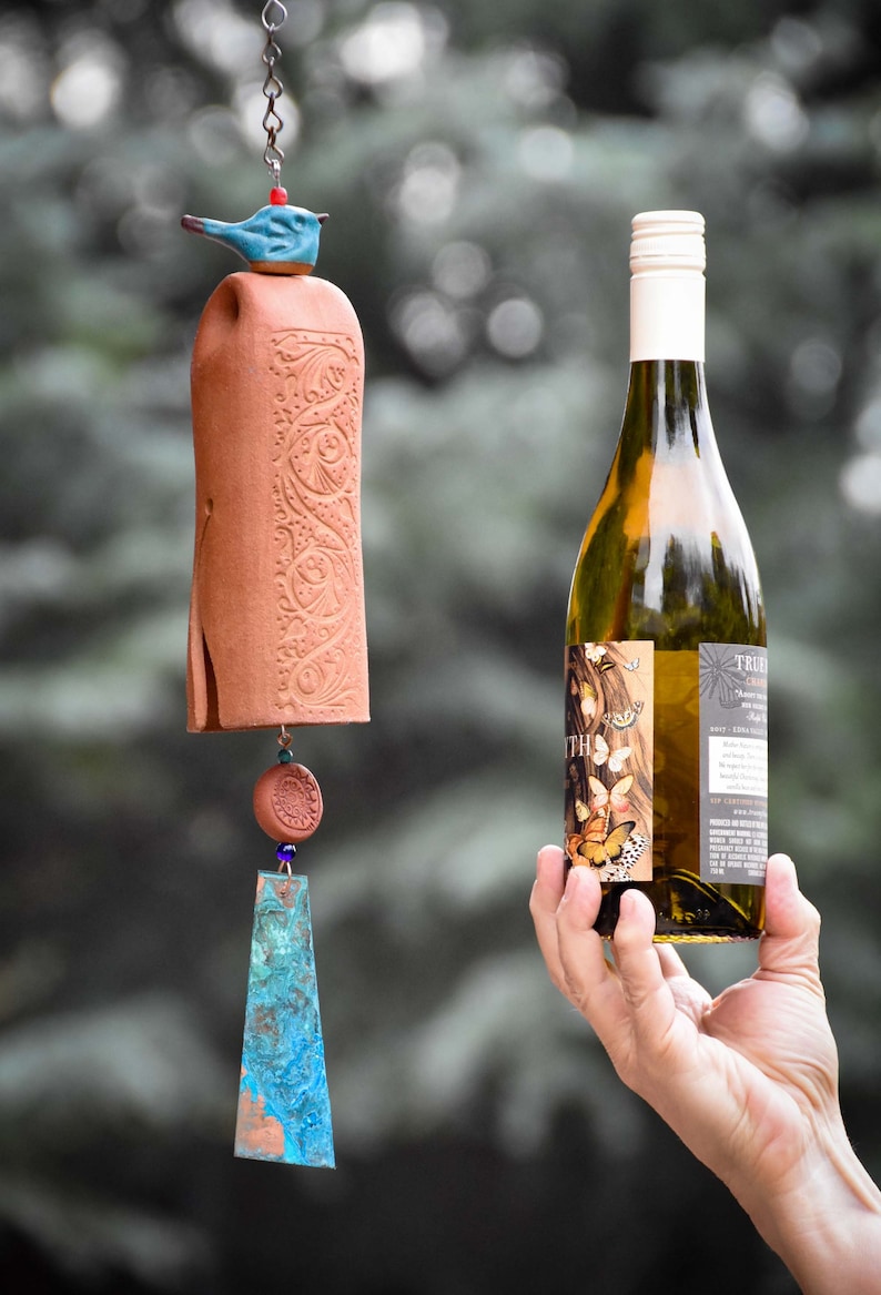 May include: A terracotta wind chime featuring a blue ceramic bird topper, a cylindrical body with embossed floral patterns, a small round terracotta bead, and a flat, triangular copper-toned metal pendant with a blue and green patina finish.