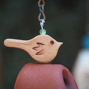 May include: A tan ceramic bird ornament with a speckled texture and carved wing detail, suspended by a metal chain with a small translucent light green bead. The bird is positioned above a smooth, curved, reddish-brown terracotta clay element.