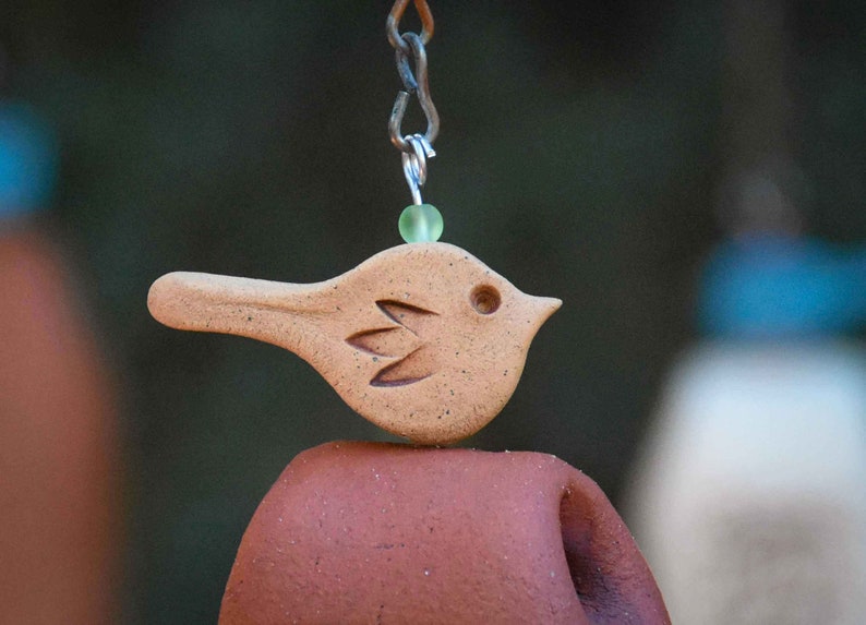 May include: A tan ceramic bird ornament with a speckled texture and carved wing detail, suspended by a metal chain with a small translucent light green bead. The bird is positioned above a smooth, curved, reddish-brown terracotta clay element.